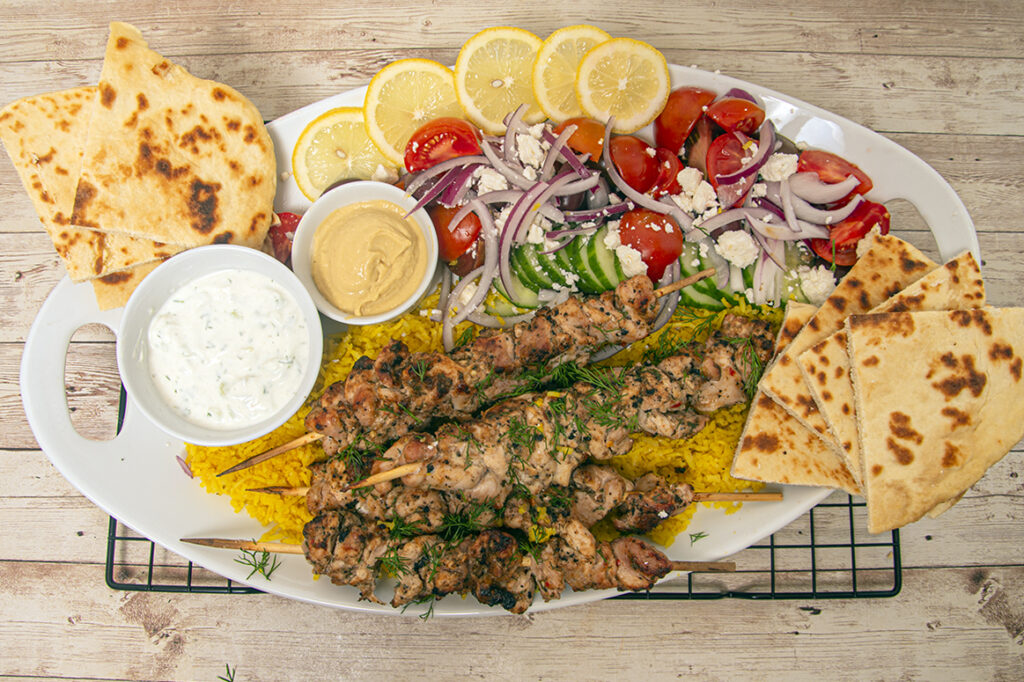 Plated Mediterranean chicken kabobs with rice and a tomato, cucumber, olive and onion salad. Topped with feta, served with naan bread, tzatziki sauce and hummus. 